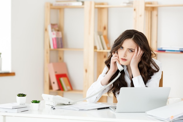 Cansado hermosa empresaria sosteniendo la mano en la cabeza mientras trabaja en la computadora y algunos documentos comerciales en la oficina brillante