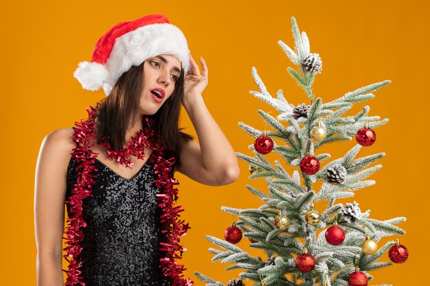 Cansado inclinando la cabeza joven hermosa niña con sombrero de navidad con guirnalda en el cuello de pie cerca del árbol de navidad poniendo la mano en la cabeza aislada sobre fondo naranja
