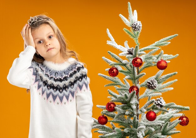 Cansado, inclinar la cabeza, niña, posición, cerca, árbol de navidad, llevando, tiara, con, garland, en, cuello, poniendo, mano, cabeza, aislado, en, fondo naranja