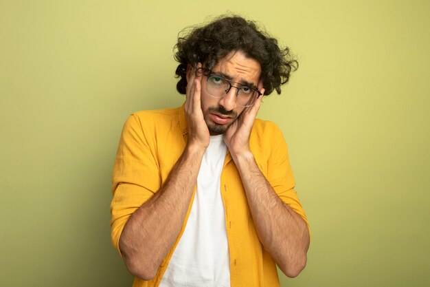 Cansado joven apuesto hombre caucásico con gafas poniendo las manos en la cabeza mirando a cámara aislada sobre fondo verde oliva con espacio de copia