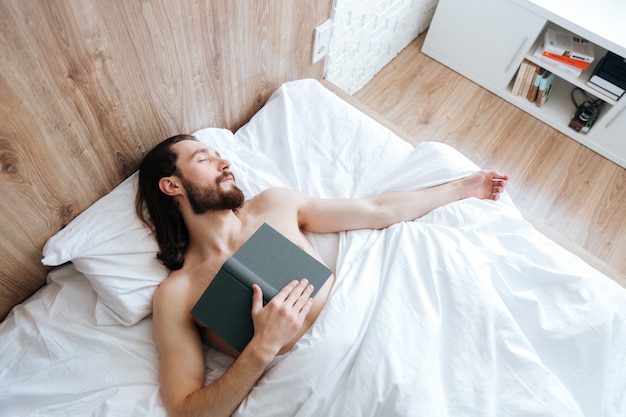 Cansado joven barbudo con libro durmiendo en la cama