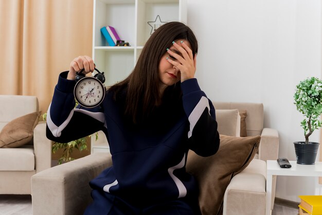 Cansado joven bastante caucásica sentado en un sillón en la sala de estar diseñada sosteniendo el reloj despertador poniendo la mano en la cara con los ojos cerrados