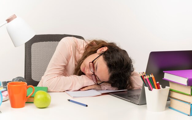 Cansado joven bonita colegiala con gafas durmiendo en un escritorio con herramientas escolares aisladas sobre fondo blanco.