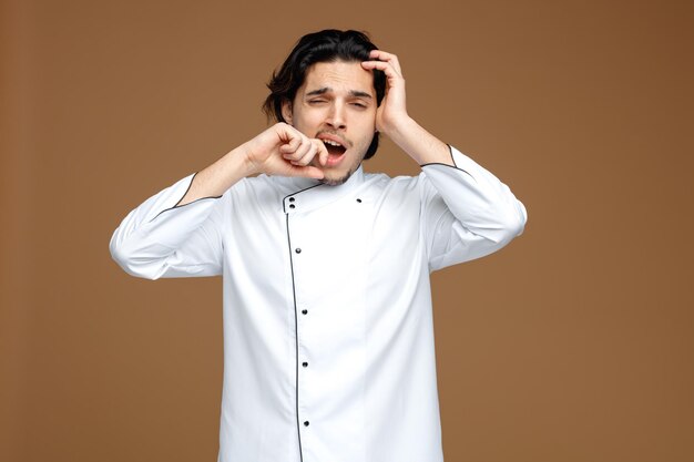 Cansado joven chef vistiendo uniforme manteniendo la mano en la cabeza tocando los labios mirando a la cámara bostezando aislado sobre fondo marrón