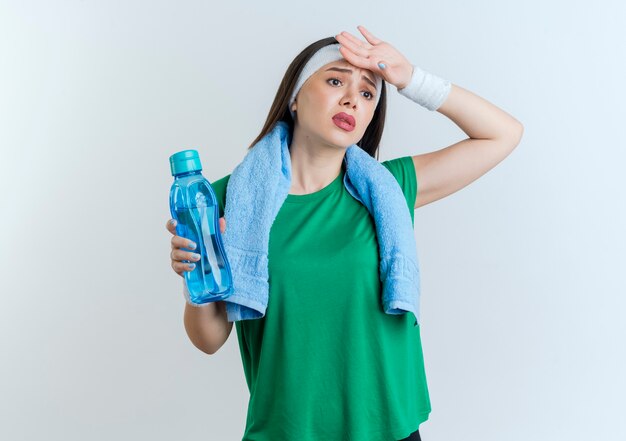 Cansado joven deportiva vistiendo diadema y muñequeras con una toalla alrededor del cuello sosteniendo una botella de agua poniendo la mano en la frente mirando de lado