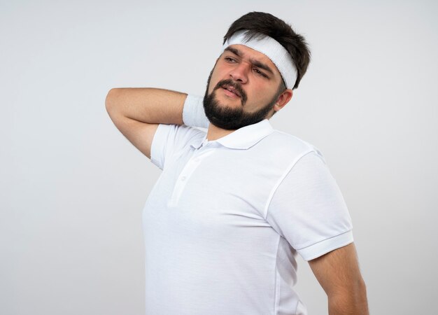 Cansado joven deportivo mirando de lado con diadema y muñequera poniendo la mano en el cuello aislado en la pared blanca