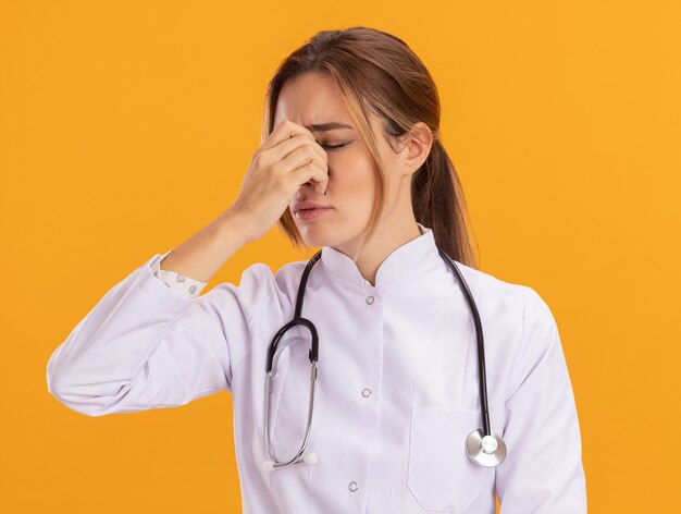 Cansado de la joven doctora cerrada con bata médica con estetoscopio agarró la nariz aislada en la pared amarilla