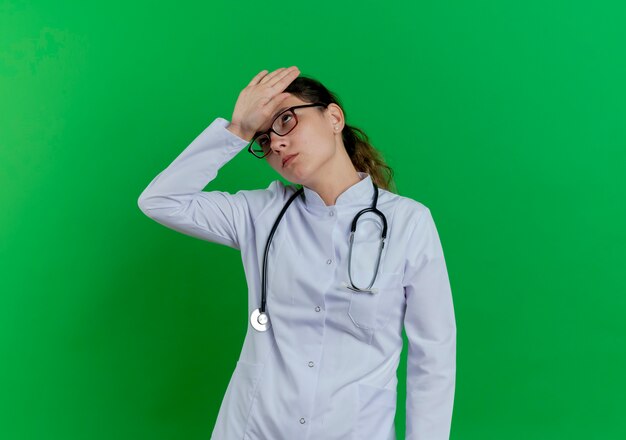 Cansado joven doctora vistiendo bata médica y estetoscopio y gafas manteniendo la mano en la frente mirando hacia arriba aislado en la pared verde con espacio de copia