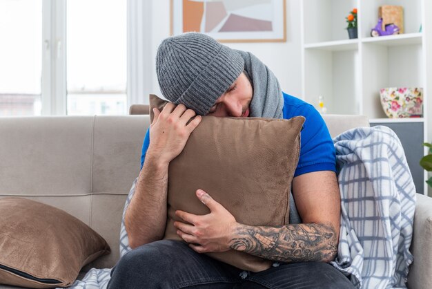 Cansado joven enfermo con bufanda y gorro de invierno sentado en el sofá en la sala de estar abrazando la almohada descansando la cabeza sobre ella con los ojos cerrados