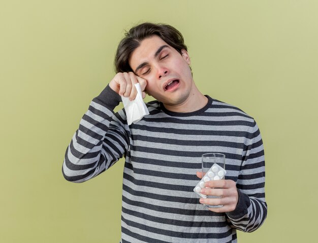 Cansado joven enfermo sosteniendo un vaso de agua con pastillas y una servilleta limpiando los ojos con la mano aislado en verde oliva