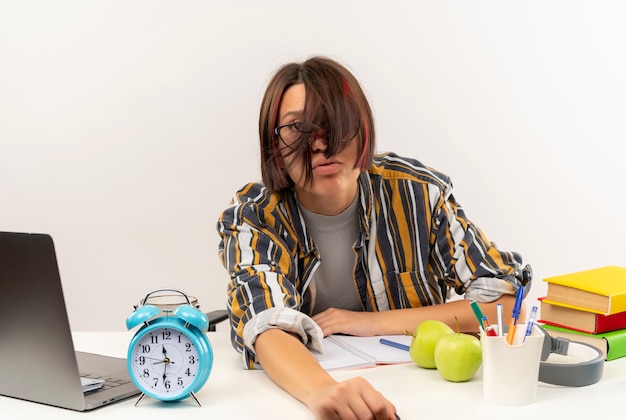 Foto gratuita cansado joven estudiante con gafas sentado en un escritorio con herramientas universitarias mirando a la cámara aislada sobre fondo blanco.