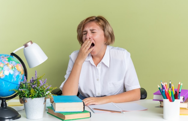 Cansado joven estudiante rubia sentada en el escritorio con herramientas escolares bostezando con los ojos cerrados aislado en la pared verde oliva