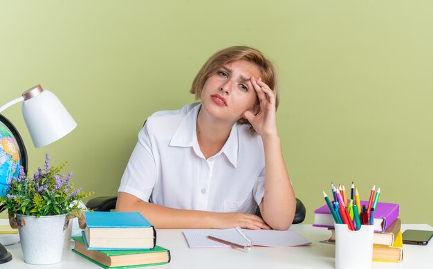 Cansado joven estudiante rubia sentada en el escritorio con herramientas escolares manteniendo la mano en la cabeza mirando a la cámara aislada en la pared verde oliva