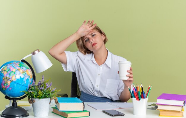 Cansado joven estudiante rubia sentada en el escritorio con herramientas escolares sosteniendo una taza de café de plástico manteniendo la mano en la frente mirando a la cámara aislada en la pared verde oliva