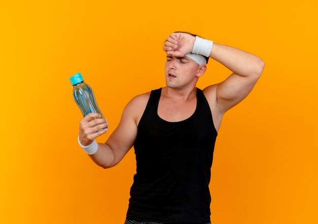 Cansado joven guapo deportivo con diadema y muñequeras sosteniendo y mirando la botella de agua con la mano en la frente aislada en la pared naranja