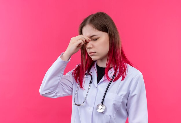 Cansado joven médico con bata médica estetoscopio poniendo los dedos en la nariz en la pared rosa aislada