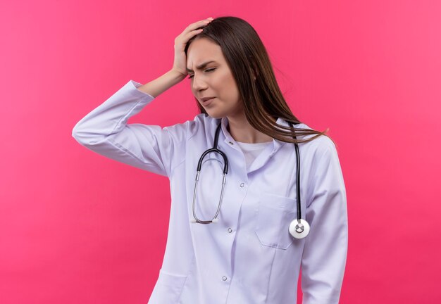 Cansado joven médico con bata médica estetoscopio puso su mano en la cabeza en la pared rosa aislada