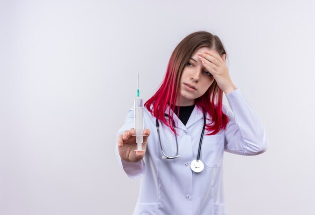 Cansado joven médico con estetoscopio bata médica mirando la jeringa en su mano macetas con la mano en la frente en la pared blanca aislada