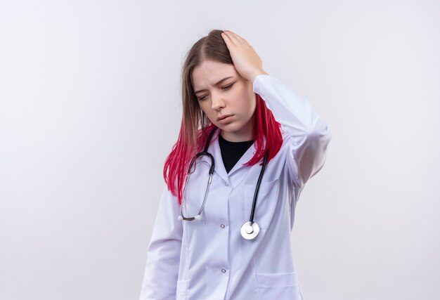 Cansado joven médico con estetoscopio bata médica poniendo la mano en la cabeza en la pared blanca aislada