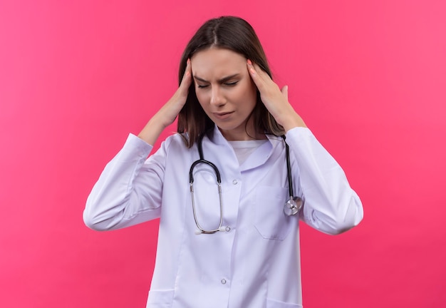 Cansado joven médico con estetoscopio bata médica puso sus manos en la frente en la pared rosa aislada