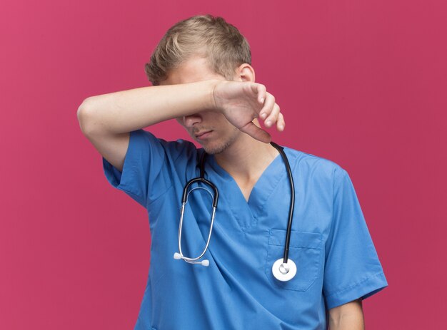 Cansado joven médico vistiendo uniforme médico con estetoscopio secándose la frente con la mano aislada en la pared rosa