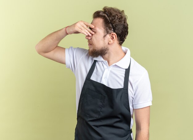 Cansado joven peluquero vistiendo uniforme limpiando los ojos con la mano aislada sobre fondo verde oliva