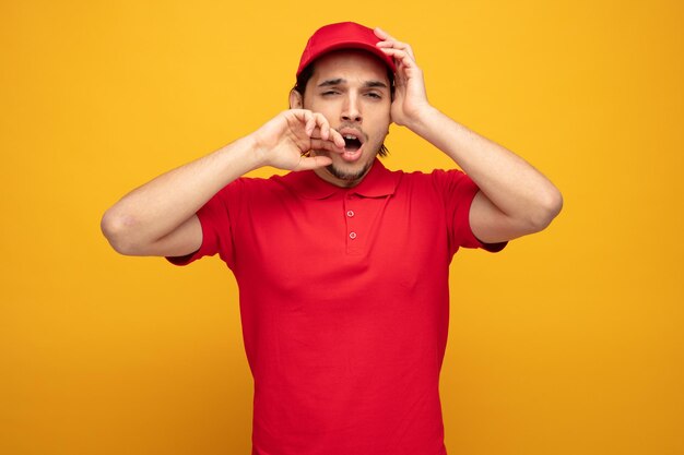 cansado joven repartidor con uniforme y gorra manteniendo la mano en la cabeza y tocando la cara mirando a la cámara bostezando aislado sobre fondo amarillo