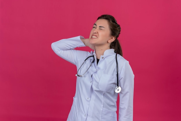 Cansado médico joven vistiendo bata médica y estetoscopio puso su mano en el cuello sobre fondo rojo isoleted