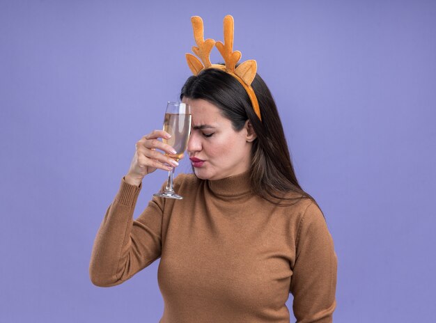 Foto gratuita cansado con los ojos cerrados hermosa joven vistiendo suéter marrón con aro de pelo de navidad sosteniendo una copa de champán en la frente aislada sobre fondo azul