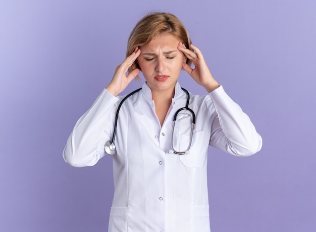 Cansado con los ojos cerrados joven doctora vistiendo bata médica con estetoscopio poniendo las manos en la sien aislado sobre fondo azul.