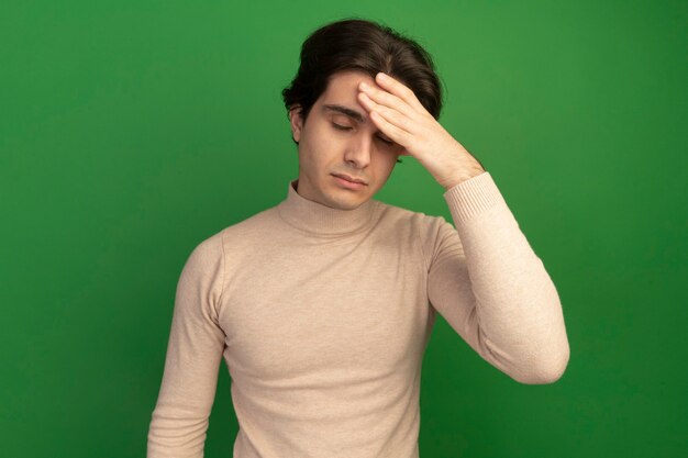 Cansado con los ojos cerrados joven guapo poniendo la mano en la frente aislada en la pared verde