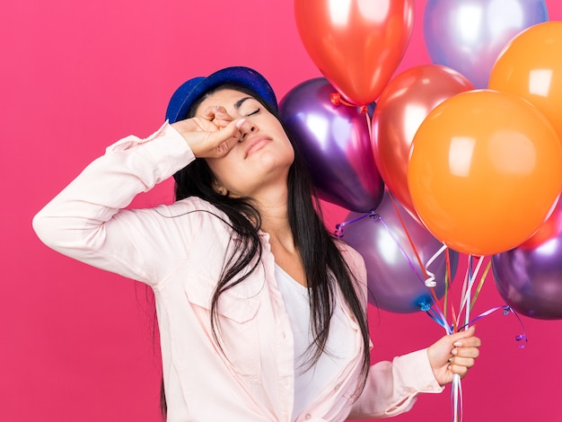 Cansado con los ojos cerrados joven hermosa mujer con sombrero de fiesta sosteniendo globos limpiando los ojos con la mano aislada en la pared rosa