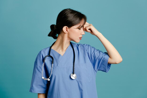 Cansado de poner la mano en la frente joven doctora vistiendo uniforme fith estetoscopio aislado sobre fondo azul.