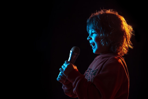 Cantando como una celebridad, estrella de rock. Retrato de niño caucásico sobre fondo oscuro de estudio en luz de neón. Preciosa modelo rizada. Concepto de emociones humanas, expresión facial, ventas, publicidad, música, hobby, sueño.