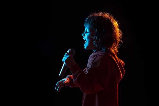 Cantando como celebridad, estrella de rock. Retrato de niño caucásico sobre fondo oscuro de estudio en luz de neón. Preciosa modelo rizada.