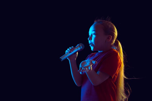 Cantando feliz. Retrato de niña caucásica en la pared oscura en luz de neón. Hermosa modelo de mujer con altavoz. Concepto de emociones humanas, expresión facial, ventas, anuncios, pasatiempos, sueños, música.