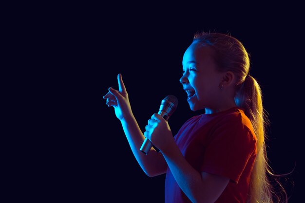 Cantando feliz. Retrato de niña caucásica sobre fondo oscuro de estudio en luz de neón. Hermosa modelo de mujer con altavoz. Concepto de emociones humanas, expresión facial, ventas, anuncios, pasatiempos, sueños, música.