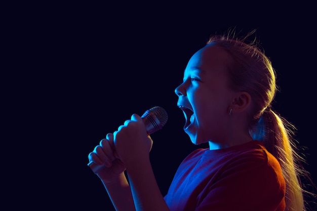 Foto gratuita cantando feliz. retrato de niña caucásica sobre fondo oscuro de estudio en luz de neón. hermosa modelo de mujer con altavoz.