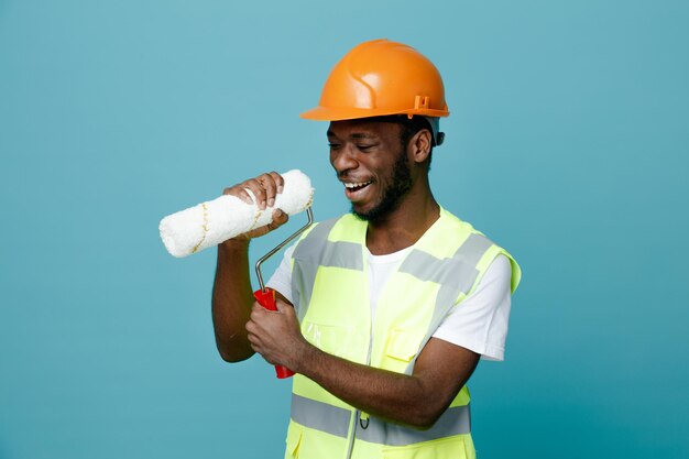 Cantando joven constructor afroamericano en uniforme sosteniendo cepillo de rodillos aislado sobre fondo azul.