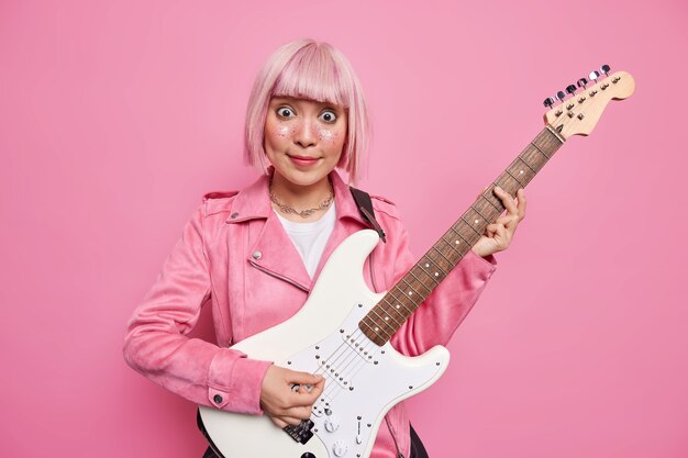 La cantante asiática sorprendida con el pelo rosado toca la guitarra eléctrica como parte de la popular banda. El talentoso músico realiza música rock en el estudio se prepara para el concierto. Estilo retro. Instrumentos musicales