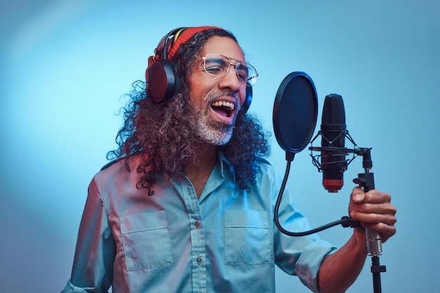 Un cantante rastafari africano que usa una camisa azul y un gorro escribe emocionalmente una canción en el estudio de grabación. Aislado sobre un fondo azul.