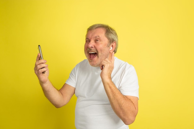 Cantar con auriculares y smartphone. Retrato de hombre caucásico sobre fondo amarillo de estudio. Hermoso modelo masculino en camisa blanca.