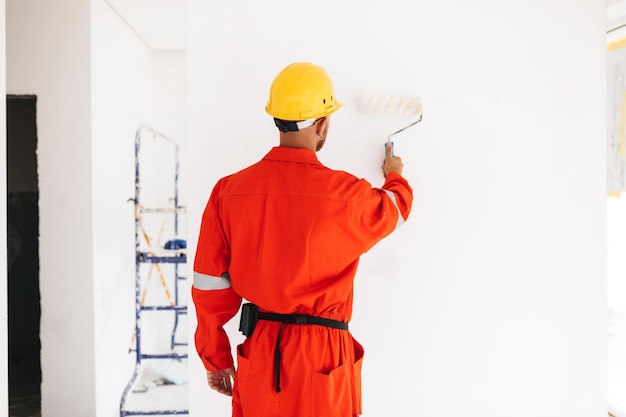 Capataz joven de pie desde atrás en ropa de trabajo naranja y casco amarillo usando rodillo de pintura en un nuevo apartamento en el trabajo