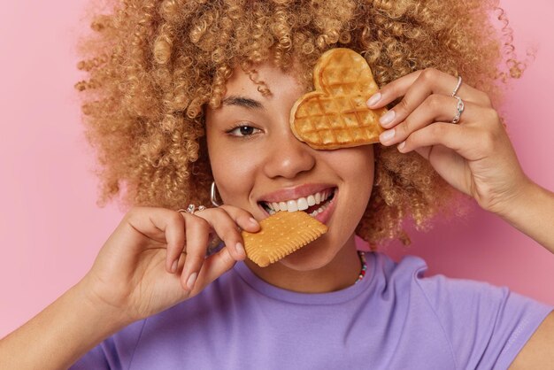 Captura de cabeza de una adolescente despreocupada y rizada que muerde galletas cubre los ojos con un gofre apetitoso tiene un gusto por lo dulce que disfruta comiendo comida deliciosa usa una camiseta púrpura casual aislada sobre un fondo rosado