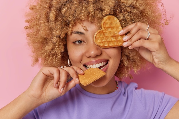 Captura de cabeza de una adolescente despreocupada y rizada que muerde galletas cubre los ojos con un gofre apetitoso tiene un gusto por lo dulce que disfruta comiendo comida deliciosa usa una camiseta púrpura casual aislada sobre un fondo rosado