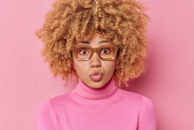 Captura de cabeza de una encantadora mujer de cabello rizado que mantiene los labios redondeados quiere besar a alguien que tiene una expresión romántica, usa anteojos y cuello de tortuga aislado sobre un fondo rosado. Modelo femenino sopla mwah.