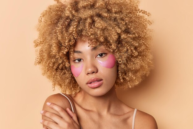 Captura de cabeza de una hermosa joven de cabello rizado que tiene una piel suave que toca el hombro mira suavemente directamente a la cámara y aplica parches de hidrogel rosados debajo de los ojos aislados sobre un fondo marrón. concepto de belleza