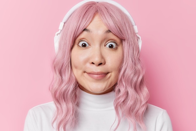 Captura de cabeza de una joven asiática con cabello rosado que se ve con los ojos muy abiertos se siente asombrada usa un cuello de tortuga blanco descubre noticias impactantes escucha una pista de audio a través de auriculares aislados sobre una pared rosada