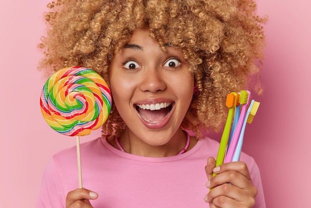 Foto gratuita captura de cabeza de una joven sorprendida de pelo rizado que mira impresionada, tiene los ojos llenos de felicidad, sostiene una gran piruleta arcoíris y los cepillos de dientes comen alimentos dañinos para los dientes aislados en un fondo rosado