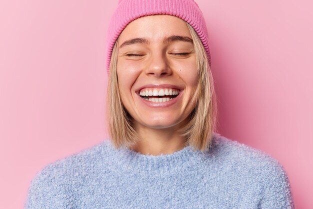 Captura de cabeza de una mujer optimista que mantiene los ojos cerrados, sonríe ampliamente, muestra dientes blancos, expresa sentimientos sinceros, se siente muy contenta, usa sombrero y un puente casual aislado sobre un fondo rosa Concepto de emociones